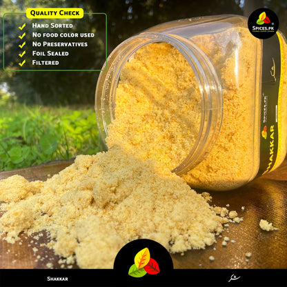 Close-up of authentic, hand-sorted golden jaggery powder spilling beautifully out of a clear plastic container.Spice container with yellow powder on a wooden surface, with 'Shakkar' branding.