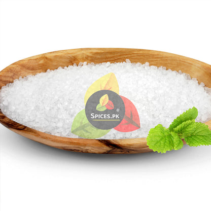 Pure white food-grade citric acid crystals (tatri) displayed in an oval wooden bowl with a fresh mint leaf by Spices.pk.