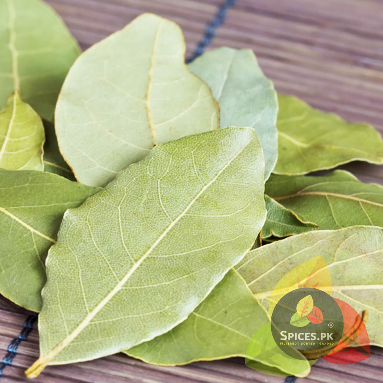 Close-up of green leaves on a wooden surface with Spices.PK logo.High-quality dried whole Bay leaves (Tezz Pata) with visible veins, perfect for aromatic Pakistani and Indian cooking.