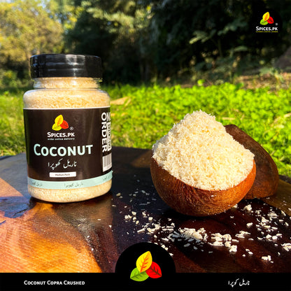 Spices.pk Khopra Crush jar displayed outdoors, emphasizing hand-sorted, chemical-free, organic shredded coconut.Jar of coconut copra crushed and a half-coconut with coconut pieces on a wooden surface outdoors.