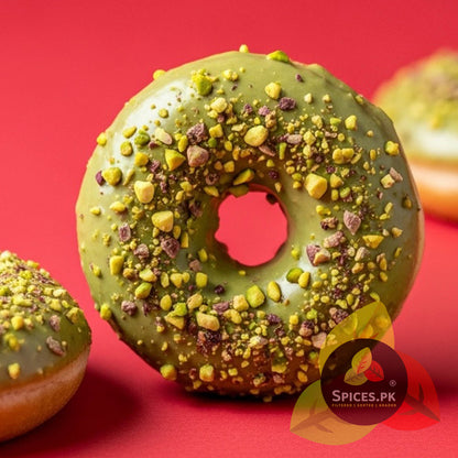 A sweet donut with green pistachio glaze, topped with crushed roasted pista pieces, demonstrating culinary uses of Spices.pk nuts.Green doughnut with pistachio pieces on a red background, branded with Spices.PK.