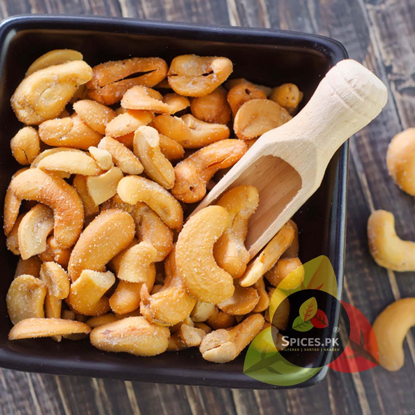 op view of high-quality crispy roasted and salted kaju filled in a black square bowl with a wooden scoop by Spices.pk.Roasted cashews in a black container with a wooden scoop on a wooden surface, branded 'Spices.Pk'.