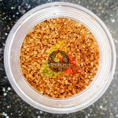 Top-down close-up view of coarse, pure brown sugar crystals with rich molasses color inside a Spices.pk jar.Container of colorful sugar with 'Spices PK' logo on a dark background