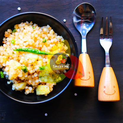 Close-up of raw sabudana pearls spilling from a wooden spoon, showcasing premium round tapioca seeds by Spices.pk.Black plate with yellow Sago dish, silver spoon, and fork with smiley face handles on a dark surface.