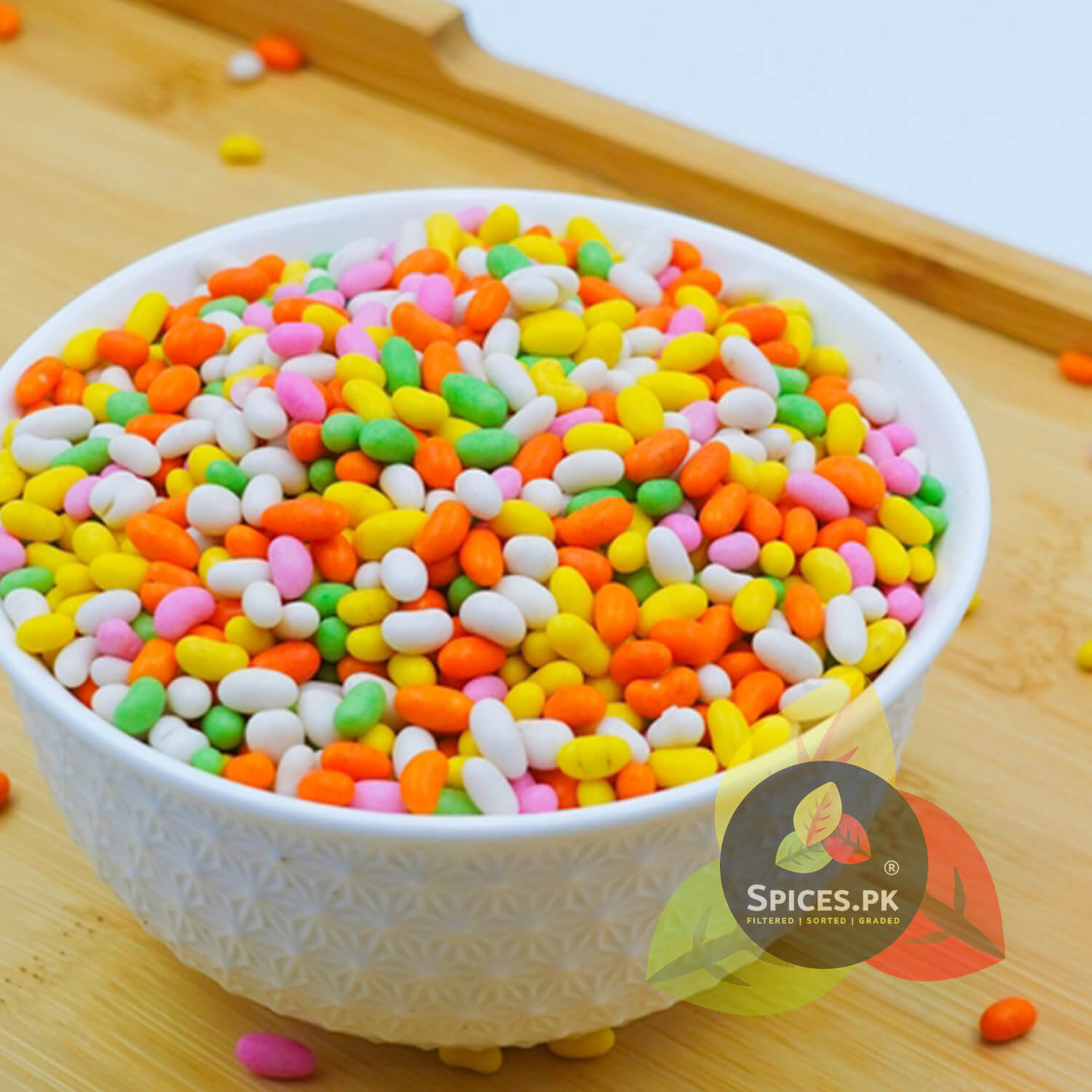 Decorative white bowl filled with multi-colored sugar saunf (sweet fennel seeds) on a wooden tray by Spices.pk.